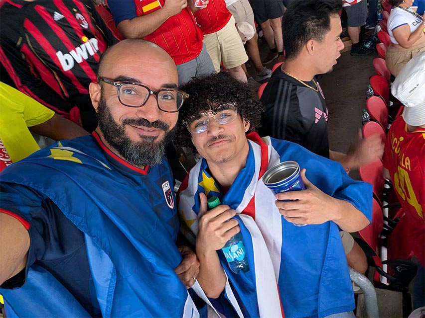 cabo verde vs spain - GC Network Solutions Two sports fans take a selfie at a stadium; one man in a blue jersey with glasses, the other with curly hair wrapped in a flag, holding a drink and a water bottle.